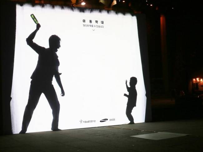 The sign displays an abusive father threatening his younger son. It is lit so that pedestrians can walk and cast their shadow on it.