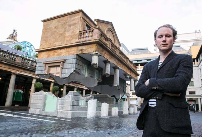 This is Alex Chinneck, pictured here with his creation. The building copies the style of the 184-year-old one behind it.